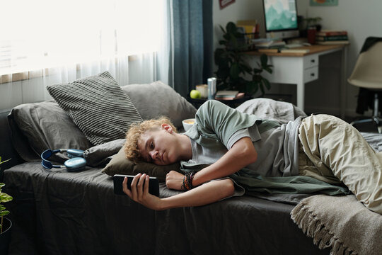 Caucasian teenage boy lying on bed holding smartphone close to face, staring at screen with tired expression, demonstrating signs of gadget addiction in bedroom setting