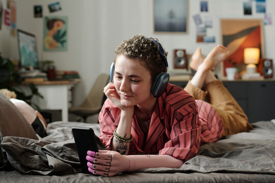Caucasian young adult woman with prosthetic arm lying on bed wearing headphones using smartphone smiling showing signs of gadget addiction in relaxed home environment - Powered by Adobe