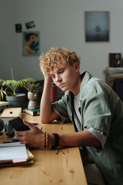 Caucasian teenage boy sitting at desk holding smartphone with bored expression resting head on hand surrounded by study materials showing signs of gadget addiction