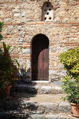 Fototapeta premium wooden door in a rustic stone wall, surrounded by green plants. steps lead up to the small archway, evoking a sense of history and secret gardens in a mediterranean setting