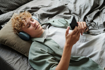 Caucasian teenage boy lying on bed wearing headphones holding smartphone close to face appearing...