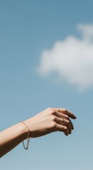 Elegant Hand Adorned with Gold Jewelry Against Serene Blue Sky and Wispy Cloud, Minimalist Outdoor Shot.