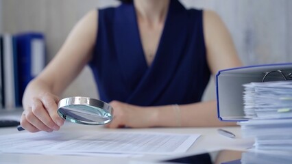 Businesswoman holding magnifying glass analyzing financial documents at office desk. Taxes, audit in business