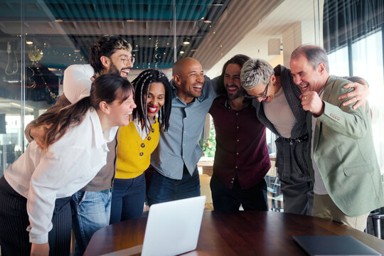 Multicultural colleagues gather in a tight huddle around a table, smiling and cheering after good news in a bright modern office, expressing unity, motivation and collaborative spirit.