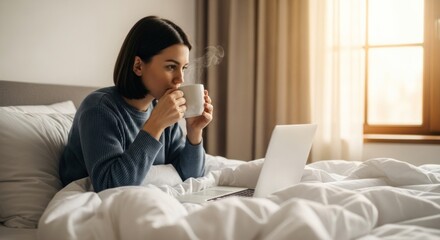 Woman sips steaming coffee in bed while working on laptop near window