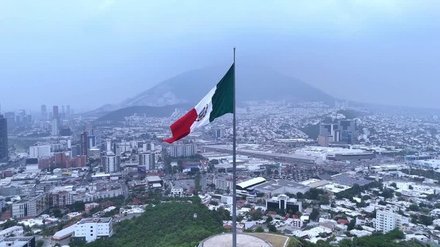 Aerial close-up of the Mexican flag waving at Cerro del Obispado, with the city of Monterrey visible in the background under a dramatic sky.