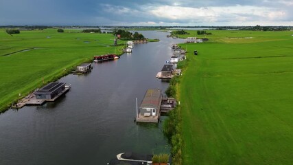 A calm aerial scene of floating houses on a river, surrounded by vast green meadows and intersecting waterways with Dutch flags gently waving in the breeze under a cloudy sky. A classic Dutch windmill