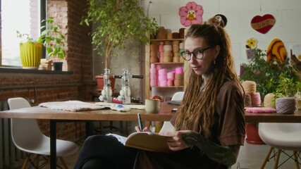 Side view shot of creative young woman drawing sketch in notebook and looking at camera with smile in rug tufting workshop, calm and cozy scene, copy space