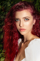 Fototapeta premium Beautiful young woman with long curly hair and gray-blue poses against the bush with hibiscus flowers. Portrait of a beautiful red-haired model with perfect skin and long eyelashes Real photo.