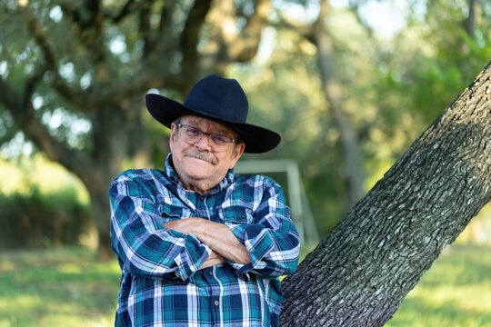 Elderly retired Caucasian man with mustache in his 80's standing outdoors with his arms crossed smiling wearing a cowboy hat looking ahead at the camera and viewer