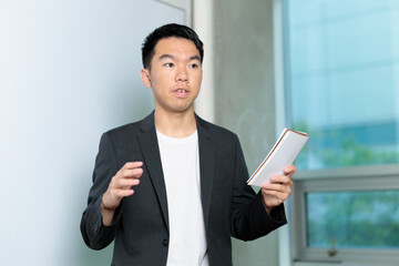 A young man speaks in front of a whiteboard while holding a notepad, presenting confidently in a classroom setting. Ideal for themes of business, education, or public speaking.