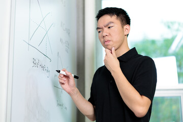 A young man stands in a classroom focused on solving a math equation on the whiteboard. He appears...