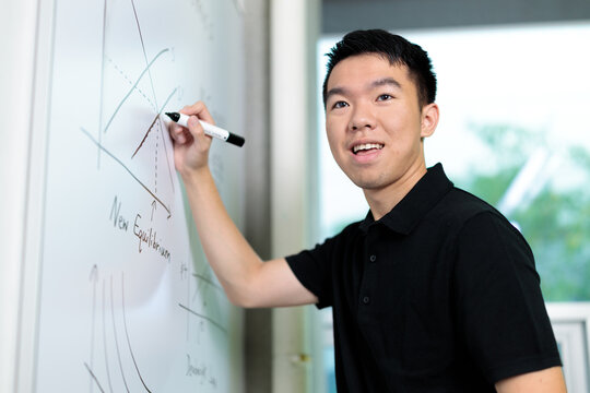 A young man writes on a whiteboard with math graphs in a university classroom. He is actively solving an academic problem during STEM class.