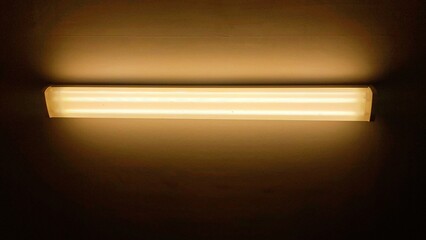 Warm amber glow from a horizontal fluorescent tube light fixture illuminating a dark, minimalist wall in a stark interior space