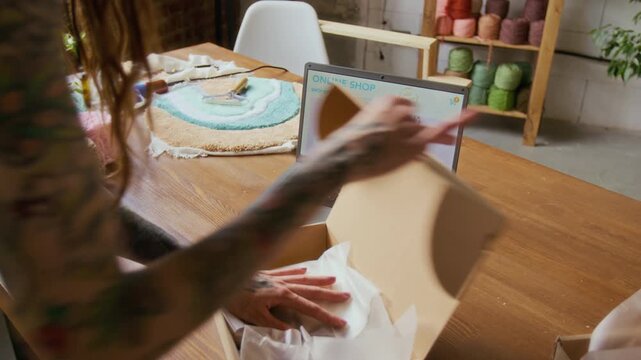 Close up shot of tattooed young woman packing small decorative rug into box while managing online shop side hustle and shipping orders, copy space