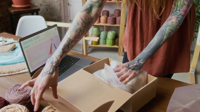 Close up shot of tattooed female hands putting small handmade rug into cardboard box while managing small business and shipping online orders, copy space