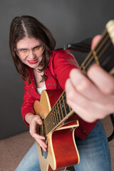 Woman playing guitar in lighthearted manner, appears to be deriving enjoyment from activity. She is doing so with smiling. Young female exudes passion and is attired in crimson leather jacket, glasses