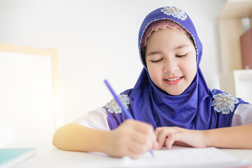 Happy Muslim girl in a purple hijab writing in her notebook at a white desk, with colorful pens beside her. Concept of education, creativity, and childhood development. she doing homework.