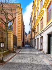 Fototapeta premium Svartmangatan, Gamla Stan—cobbled alley lined with historic brick and ocher facades in Stockholm Old Town under blue sky, a classic Europe travel scene.