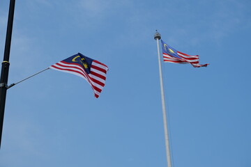 Two Malaysian national flags are waving on tall poles under a clear blue sky, symbolizing patriotism, unity, and national identity.
