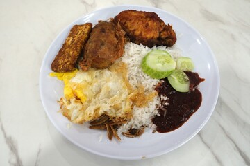 Traditional Malaysian nasi lemak served with steamed rice, fried egg, tempeh, fried chicken, cucumber slices, and spicy sambal on a white plate.
