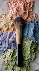 Makeup brush on a marble surface surrounded by colorful powders in orange, yellow, blue, purple and green hues, viewed from directly above