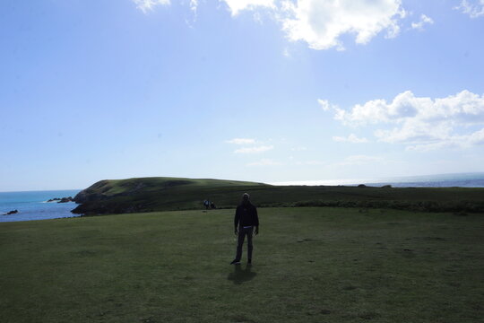 Man walking across wide grassy coastal field with ocean and headland in background under bright sky, expansive travel landscape with copy space, freedom concept and peaceful outdoor lifestyle in remot