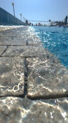 Low-angle view of a pool's edge with sunlit water and blurred figures in the background, capturing a sense of summer relaxation and refreshing vibes