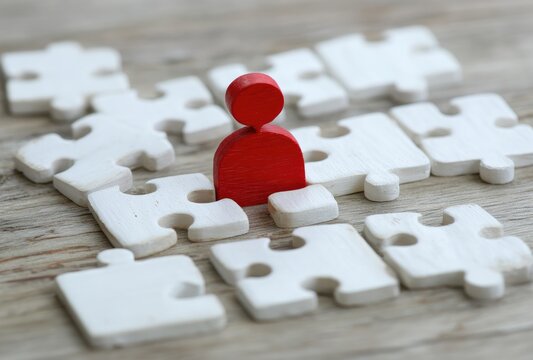 Puzzle & Red Figure A red wooden figure stands amidst unplaced white puzzle pieces on a wood grain surface, suggesting strategy or isolation