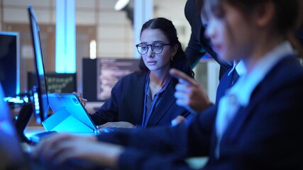 A team of data analysts and software engineers collaborates in a command center, reviewing real time analytics on a tablet while monitoring backend server performance and security logs. - Powered by Adobe