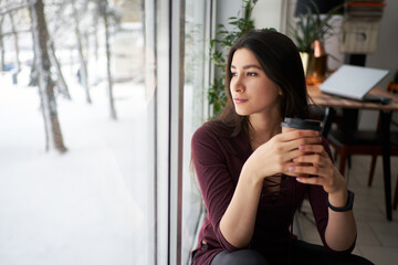 Thoughtful young asian brunette woman with cup of coffee looking through the window in winter
