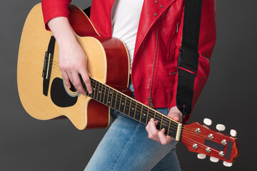 Unrecognizable woman guitarist playing acoustic guitar with concentration. Cropped view of midsection of stylish female rock musician dressed in red leather jacket and blue jeans