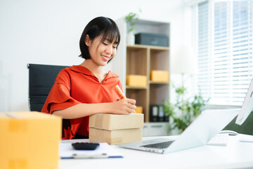 Young Asian entrepreneur writing on box in SME office with laptop and clipboard. Ideal for...