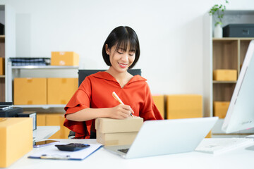 Young Asian entrepreneur writing on box in SME office with laptop and clipboard. Ideal for...