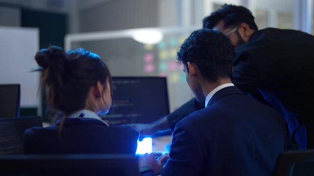 A manager trains his team in a modern office, pointing at the computer screen. The group of young professionals works together late at night to solve a difficult problem.