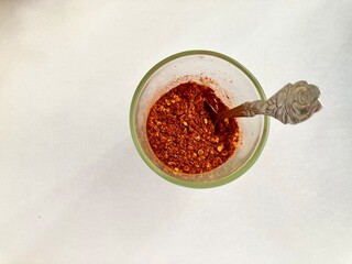 top view shot of a clear glass cup of red ้hot chili powder and a rose-patterned spoon sit on a white table with the copy space on the right side of frame