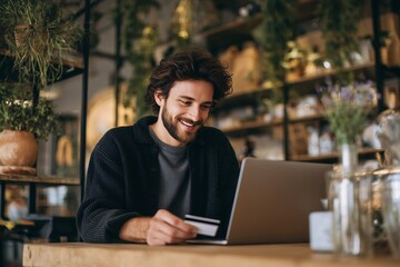 Smiling man holding a credit card while using a laptop in a cozy cafe setting, concept for online shopping, digital payments, and financial technology