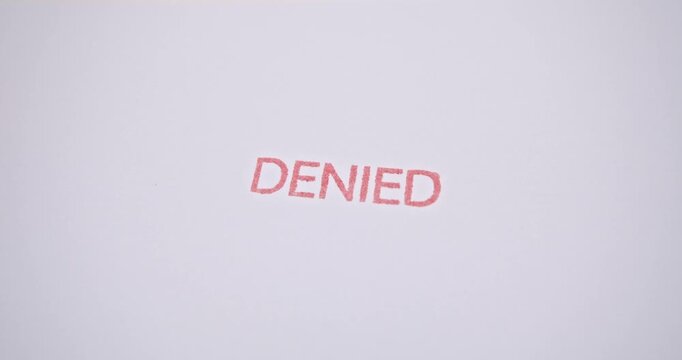 A real video frame showing a hand stamping the word &lsquo;DENIED&rsquo; in red ink on a white paper document, symbolizing the official rejection of an application, loan, visa, claim, or request.