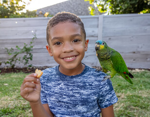A smiling young boy with a parrot perched on his shoulder enjoys a snack outdoors in a grassy backyard, showcasing a happy and friendly moment with his pet bird