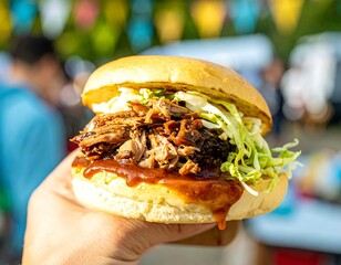 A person holds a large pulled pork sandwich with shredded lettuce and barbecue sauce in soft focus outdoor setting