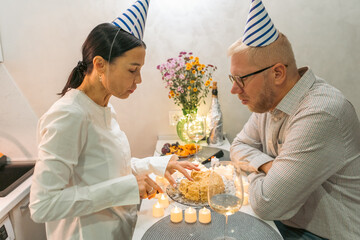 Birthday Cake Couple Celebration: Indoor intimate birthday celebration; man and woman with party hats making a wish on a cake.