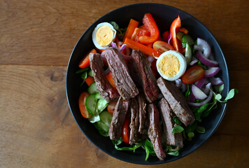 Close-up of a healthy steak salad served in a black bowl. The dish contains grilled beef slices, boiled eggs, fresh cucumber, cherry tomatoes, red onions, carrots, and bell peppers. 