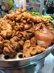 Braised pork intestine and pork trotter served on metal tray, savory street food pile with greens, glistening brown texture and aromatic appeal in busy market scene