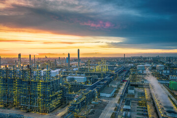 Panoramic view of a huge, illuminated petrochemical refinery complex glowing under a spectacular and dramatic sunset sky.
