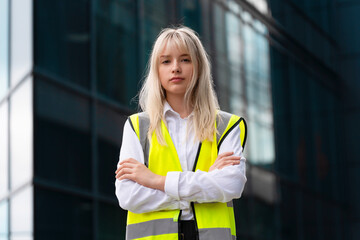 Confident construction site worker stands with arms crossed in bright safety vest against modern building backdrop