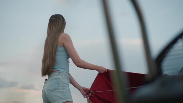 young woman in denim shorts holds red ripstop panel tether of colorful hot air balloon envelope while nearby engine blurs in foreground during breezy sunset field preflight setup scene