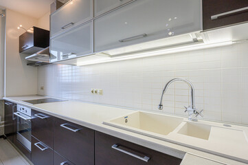 modern kitchen features dark brown cabinets, a light countertop with a double sink, and white tiled backsplash. Overhead cabinets are a mix of frosted glass and dark finishes
