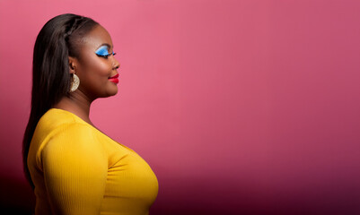 Beautiful plus size black model in makeup, wearing a yellow dress, posing against a pink background with copy space. 