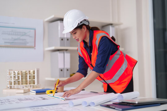Asian adult woman architect designs building plans at office desk blueprint work professional engineering project development construction safety wear