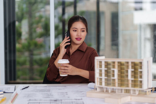 Asian Architect Professional Discusses Building Project Designs on Phone with Coffee in Modern Office Overseeing Architectural Development Planning